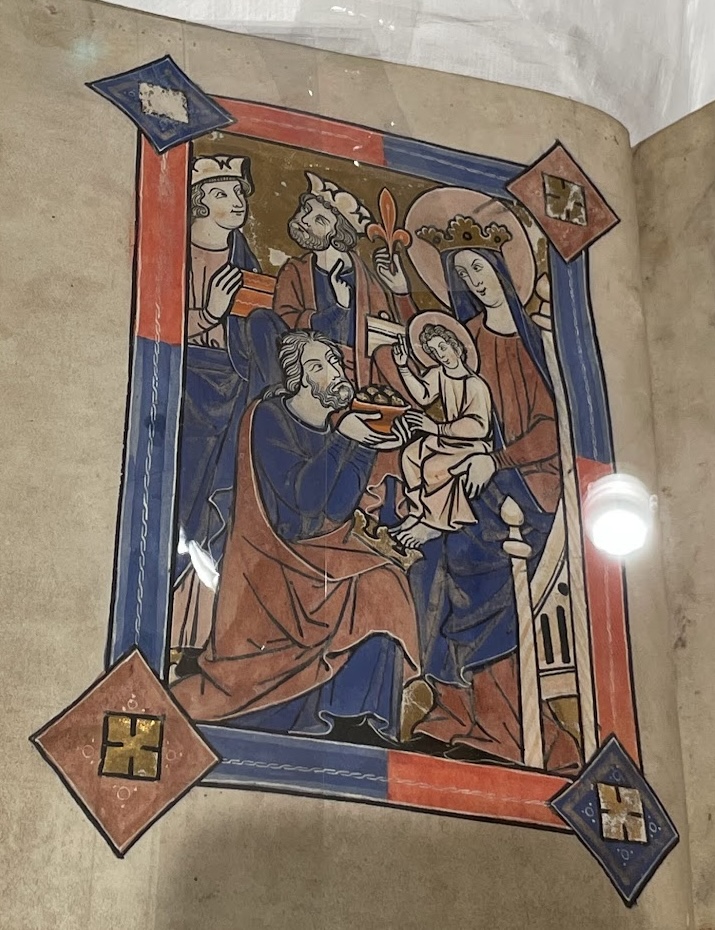 close-up on a thirteenth century psalter (MS F.4.7), open on a full page illustration of the Adoration of the Magi in shades of red and blue: the Virgin Mary, crowned with the infant Jesus on the right, holds up a red fleur-des-lys. The three kings are gathered presenting gifts, with one kneeling, his crown placed on his knee